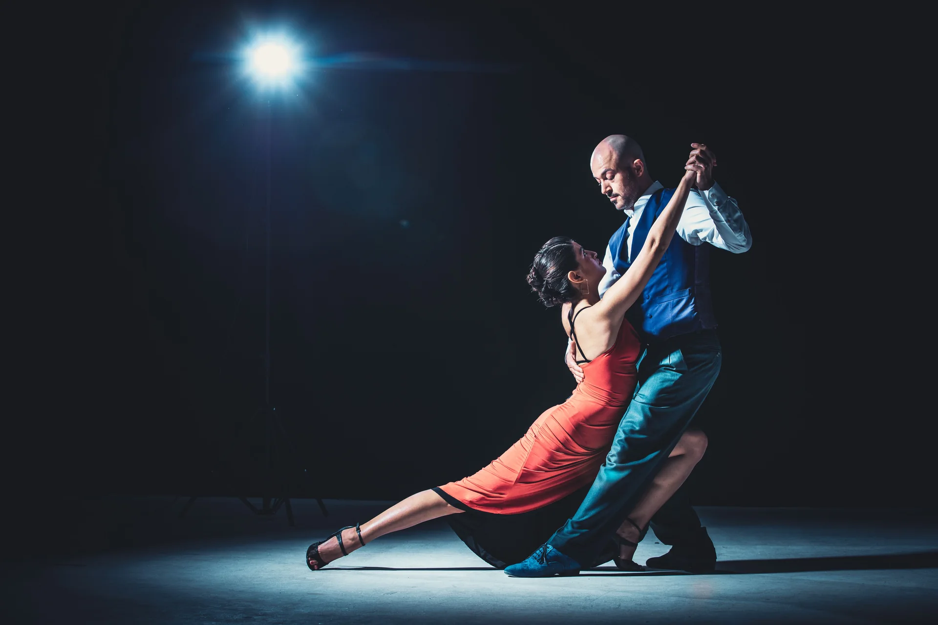 Tango pár uprostřed taneční figury v jediném světelném kuželu na tmavém jevišti, muž ve vestě a žena v červených šatech (ilustrační fotografie tanečního představení tanga, nejde o skutečný sál Señor Tango)